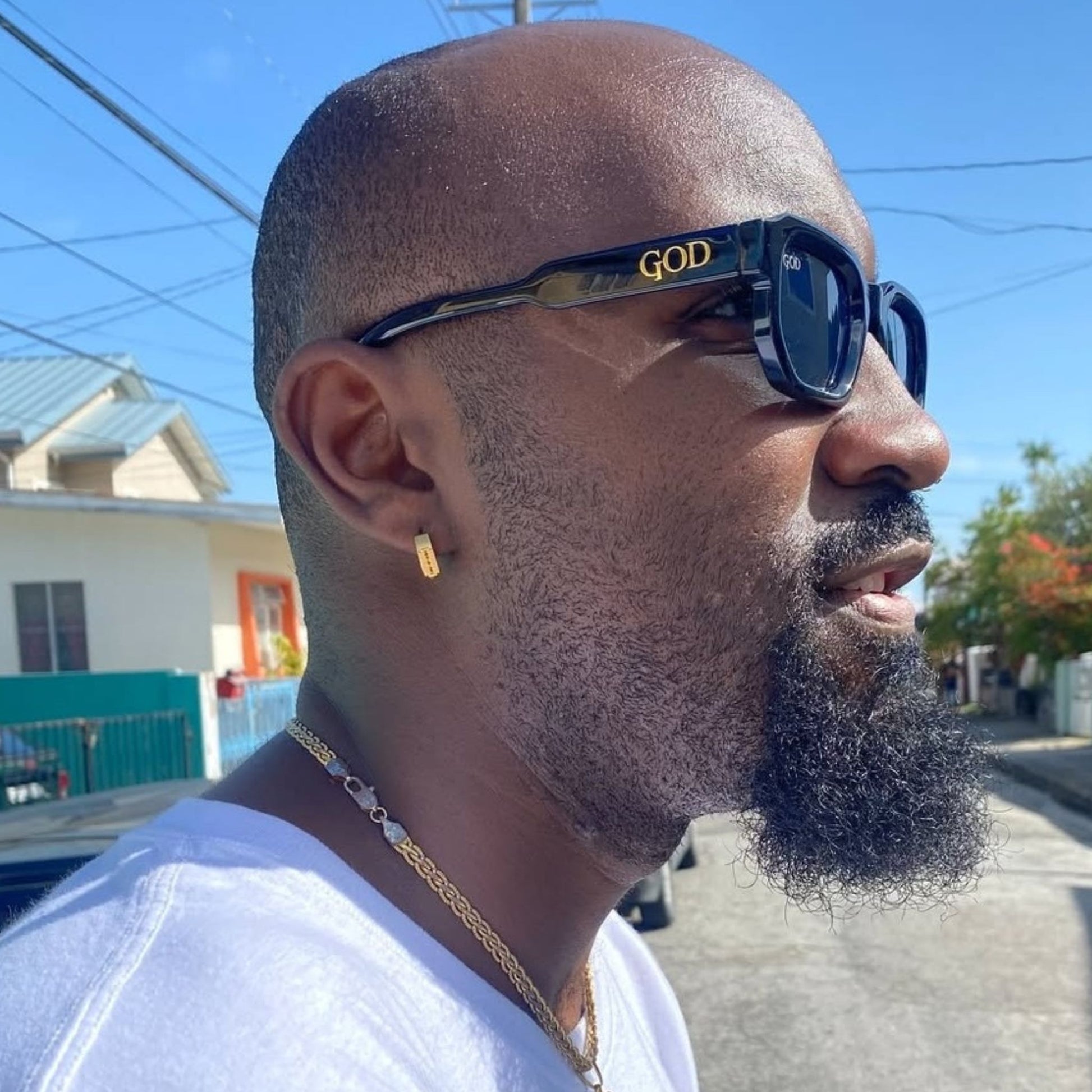 Man wearing Island God black square sunglasses with gold logo on side, standing outdoors in Caribbean sunlight.