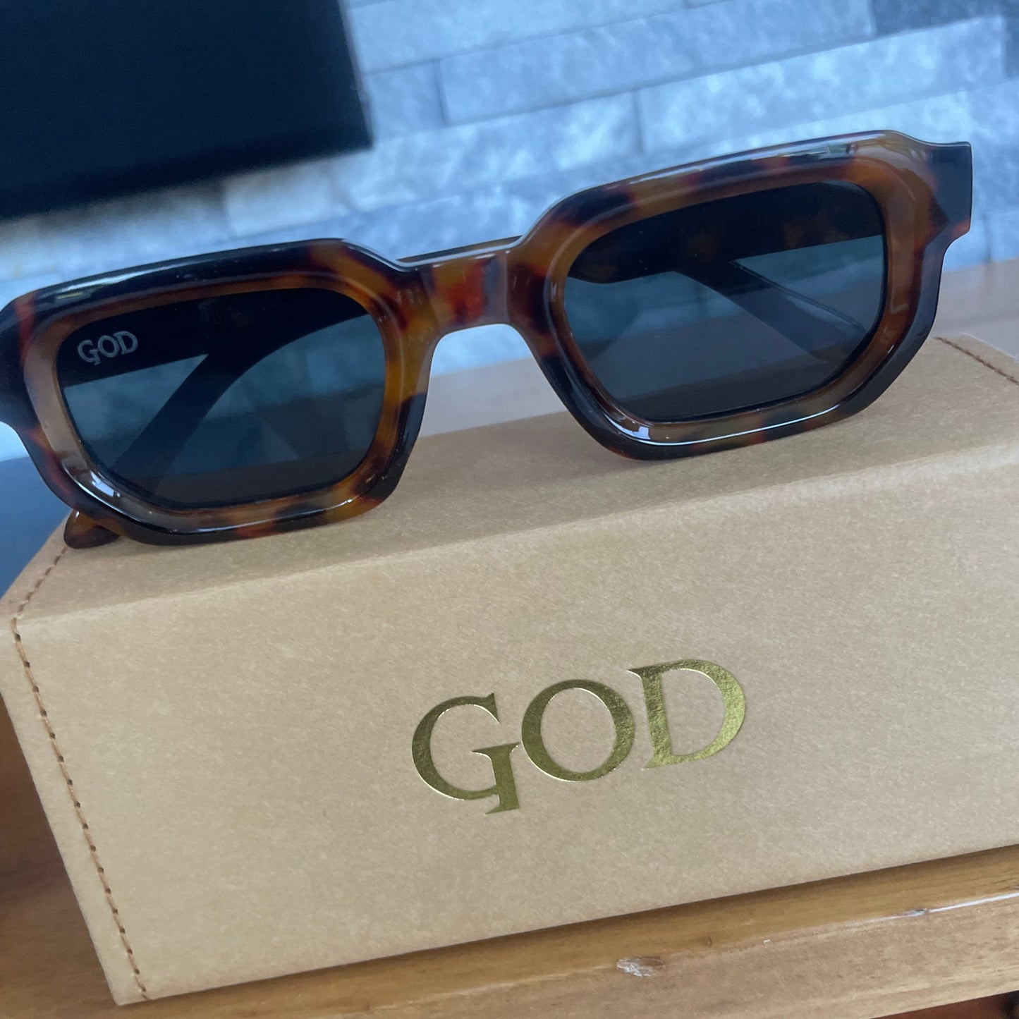 Island God brown tortoiseshell chunky rectangle sunglasses with dark lenses and gold logo, displayed on tan branded box against modern tiled background.
