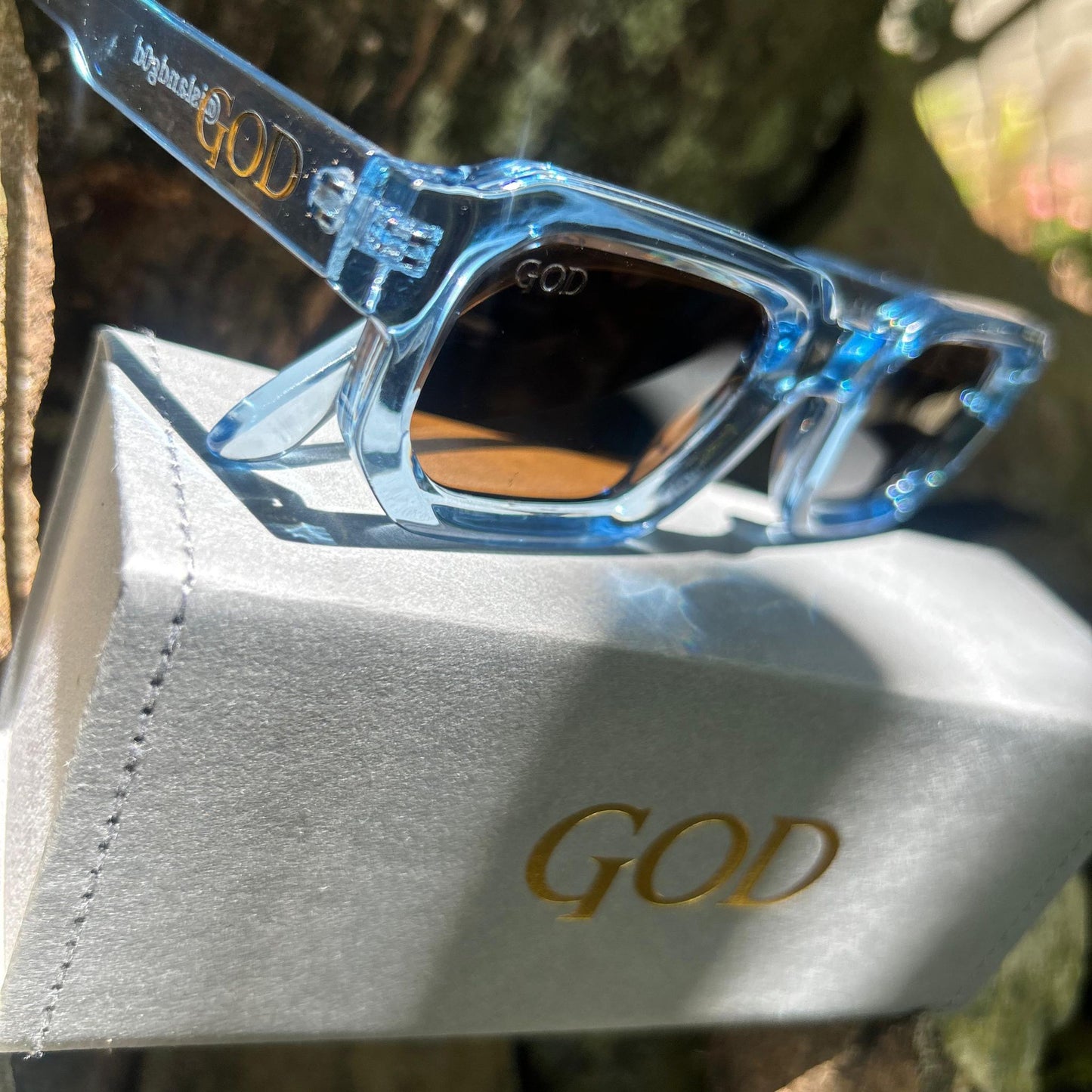 Island God clear blue chunky rectangle sunglasses with amber-tinted lenses and gold logo, displayed on grey branded case in natural outdoor setting.