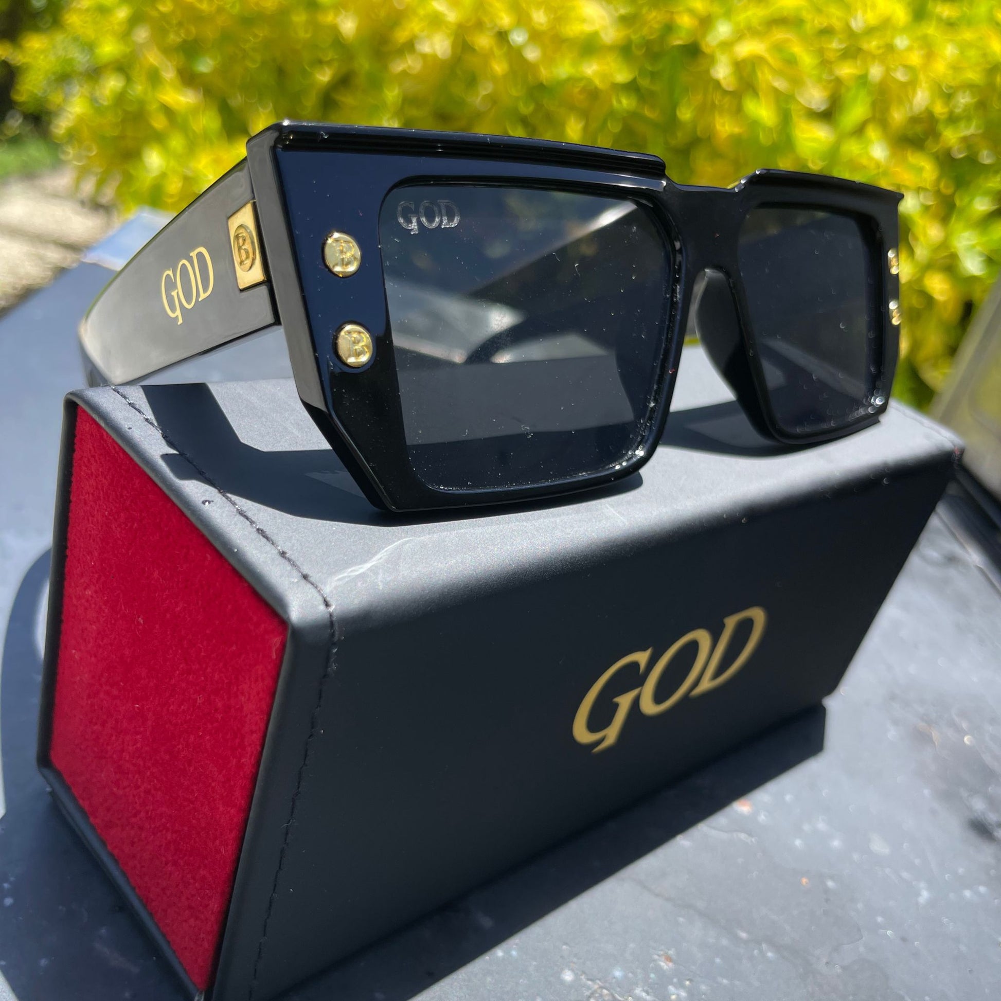 Island God black flat-top sunglasses with gold logo and stud accents, displayed on premium black and red case outdoors in natural sunlight.