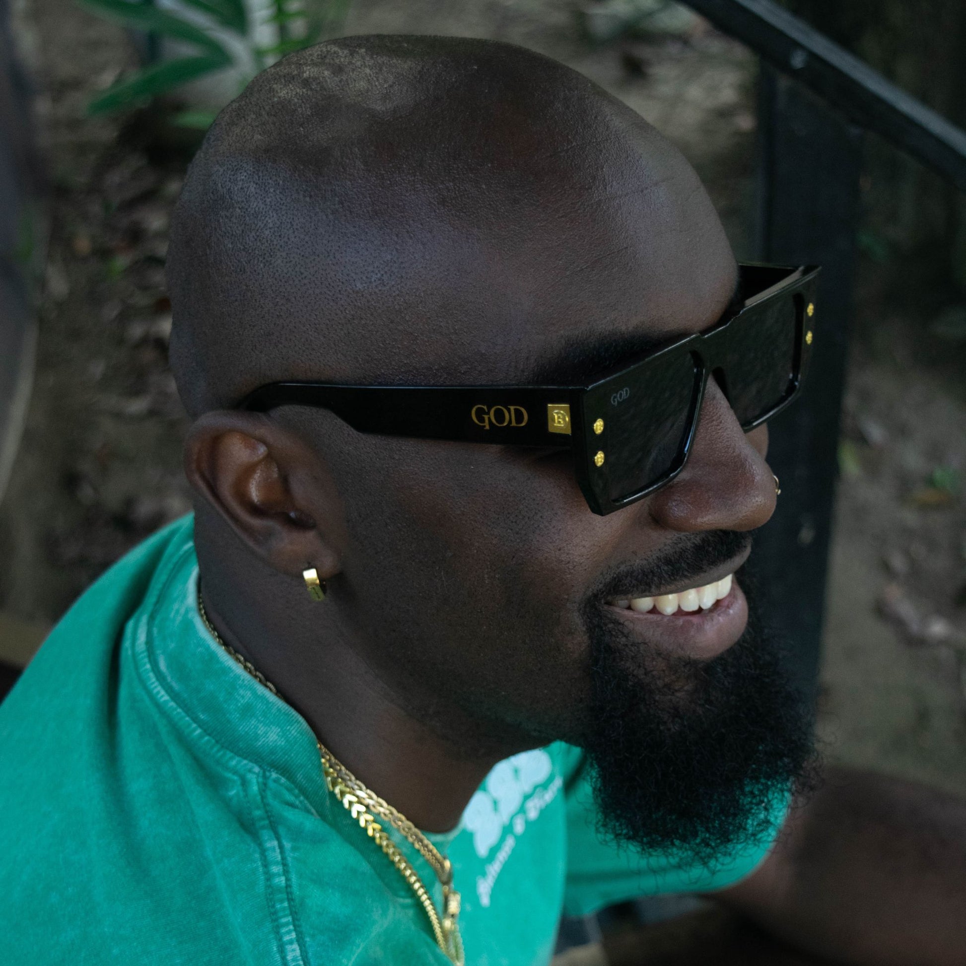 Man wearing black flat-top Island God sunglasses with gold accents and logo, styled with gold jewelry and a green shirt, smiling outdoors.