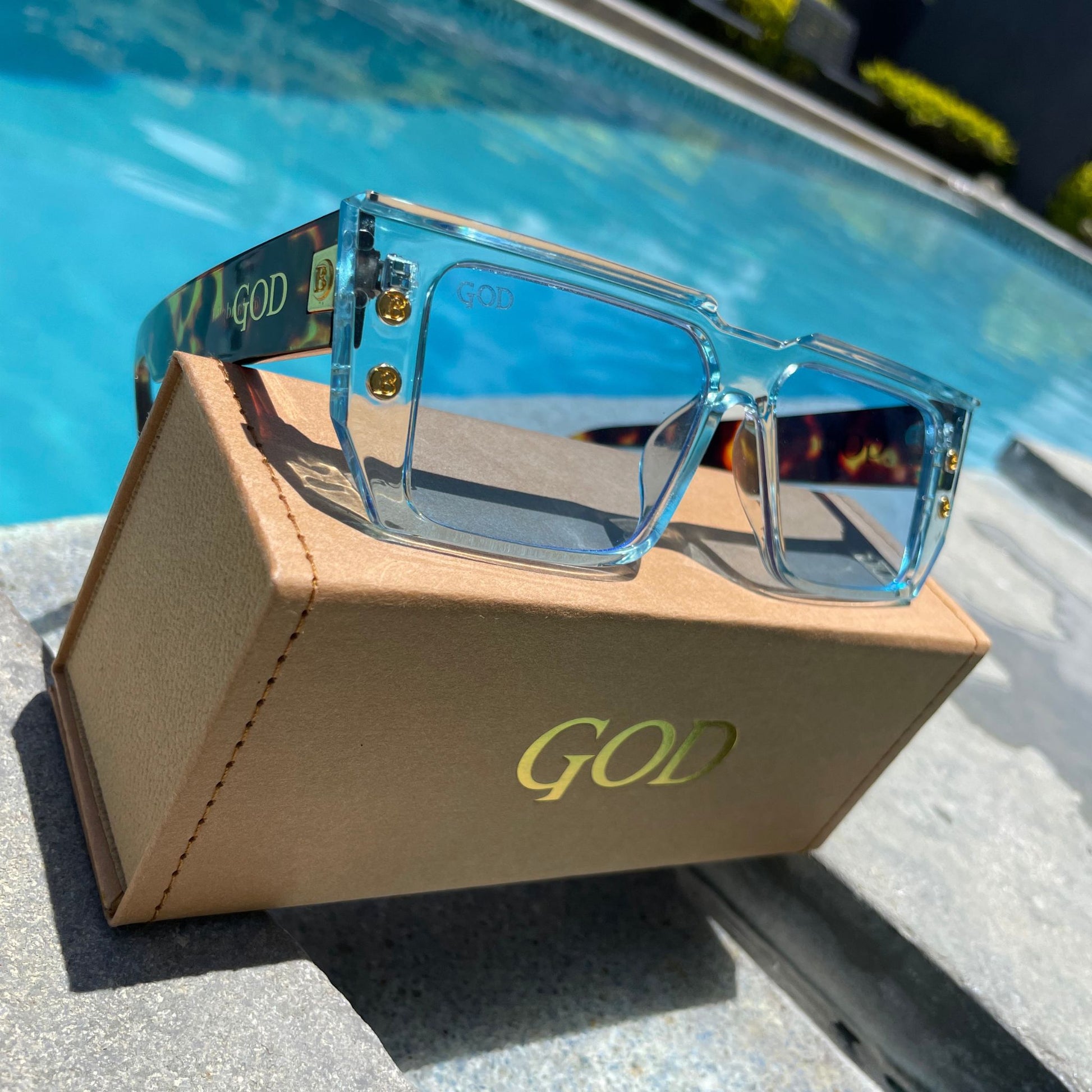 Island God flat-top blue sunglasses with gold stud detailing displayed on a tan branded case by a swimming pool, showcasing bold Caribbean-inspired eyewear style.