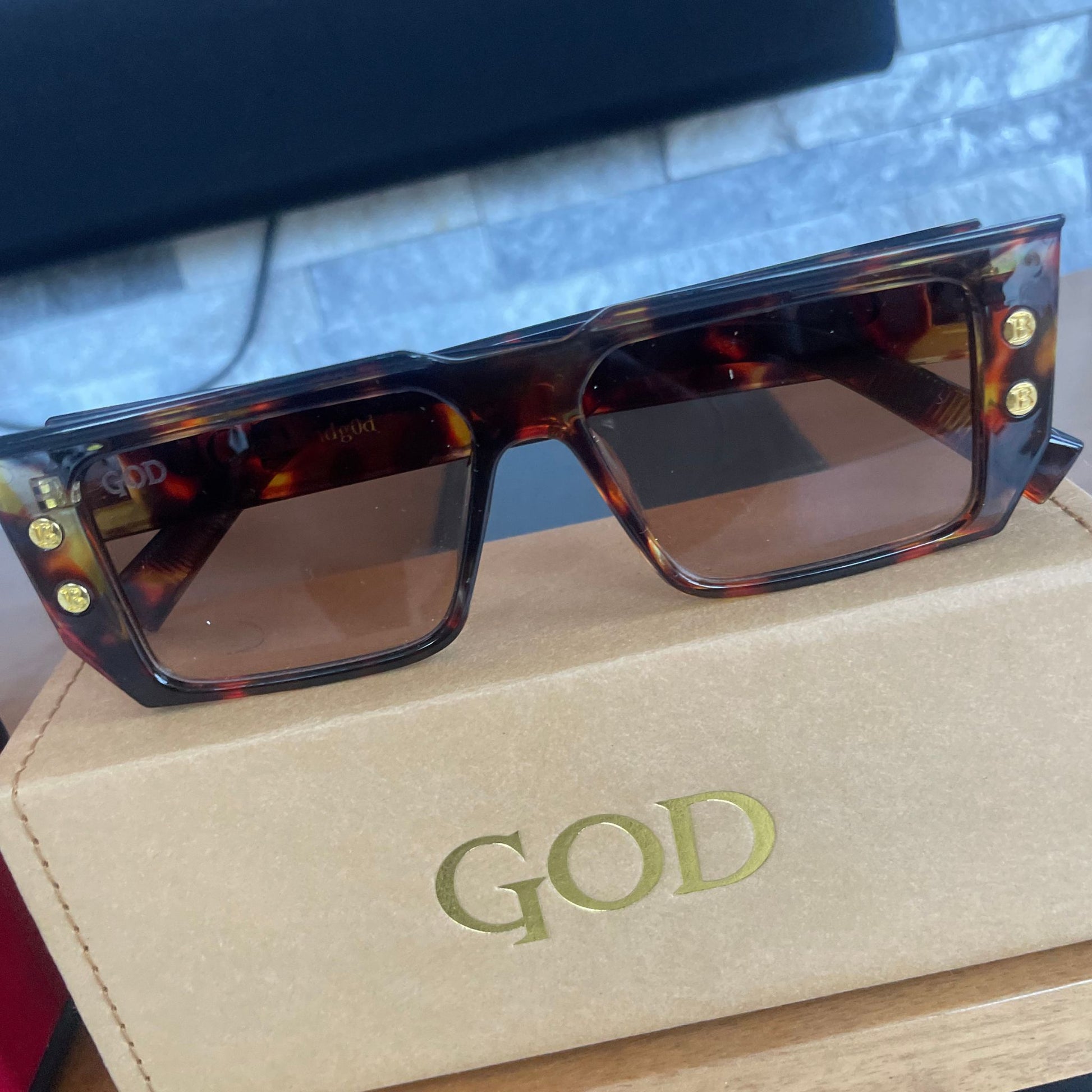 Brown Tortoiseshell flat-top Island God sunglasses with brown lenses displayed on a tan logo box, styled against a modern stone and coral background.