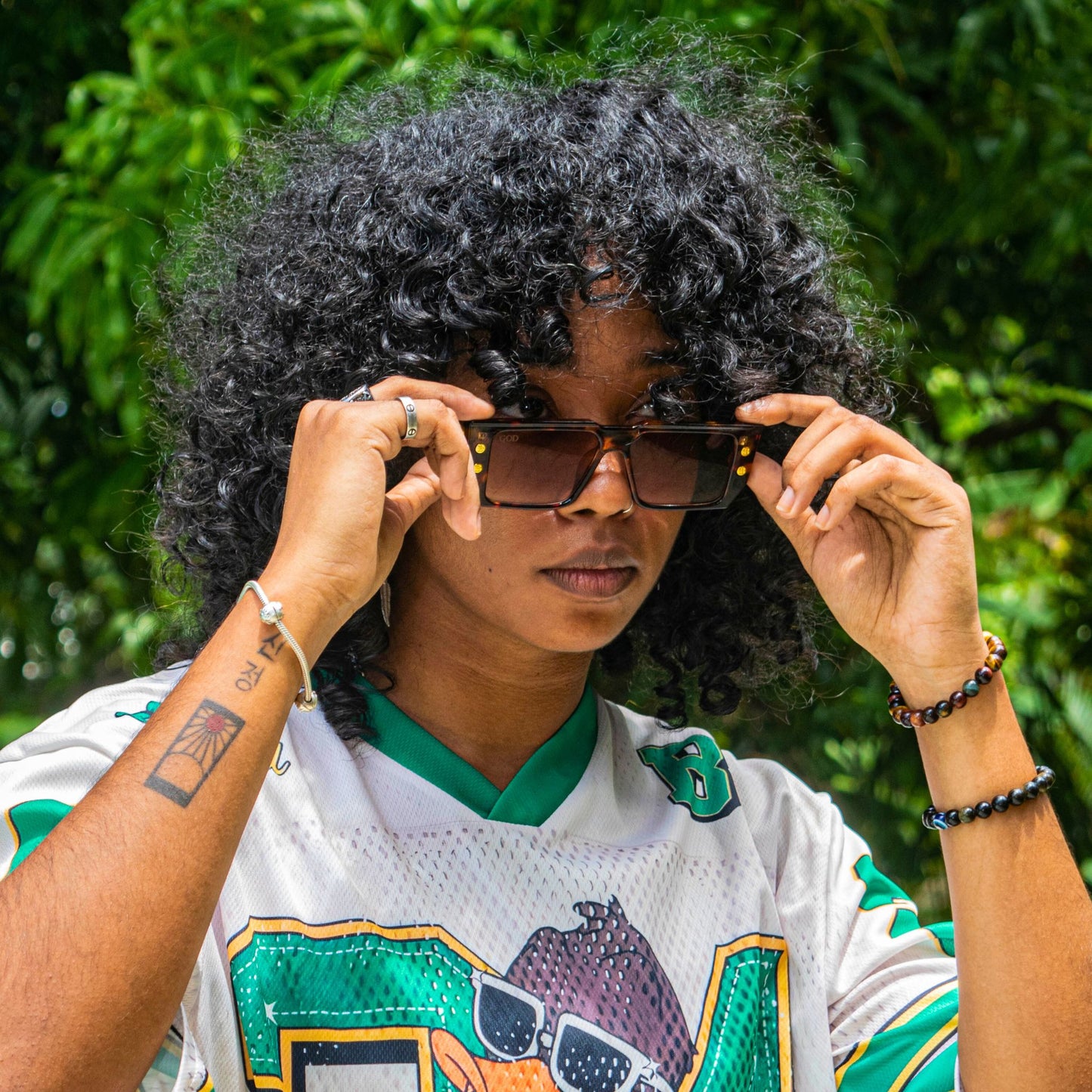 Person with curly hair wearing brown-tinted Island God flat-top sunglasses with gold accents, styled with a vintage sports jersey in a tropical outdoor setting.